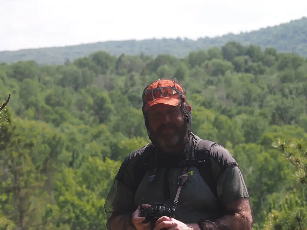 Jeff Priest smiles while holding his camera and wearing a bug jacket at a lookout in Renfrew County